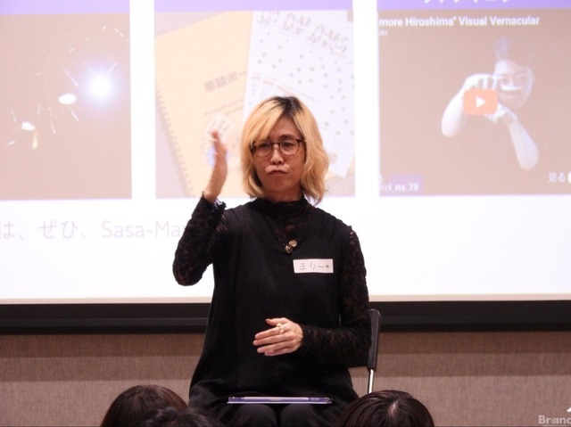 Sasa-Marieさんがスクリーンの前で手話をしている、『まりー』という名札をつけ、眼鏡をかけた金髪の人物