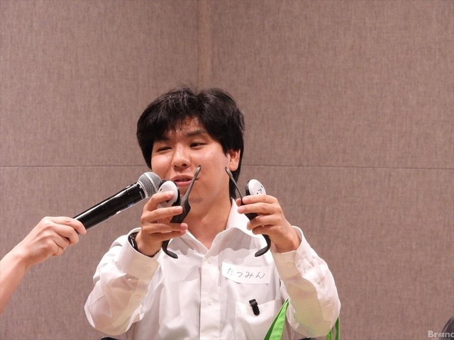 『たつみん』という名札をつけた男性の鈴木龍弥さんが、両手に黒いあしらせを持ち、差し出されたマイクに向かって笑顔で話している様子