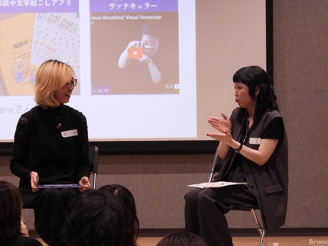 スクリーンの前で向かい合って対談する2人の女性。左側は眼鏡をかけた金髪のささまりーさん、右側は身振り手振りを交えて話す黒髪のREINAさん。背後のスクリーンには手話の動画などが映し出されている